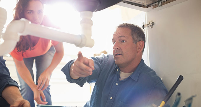 a plumber pointing to a sink drainage pipe and pointing out the problem to a female client